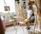 Young male artist in white shirt stained with paint sketching on easel in bright art studio with colorful painting in background.